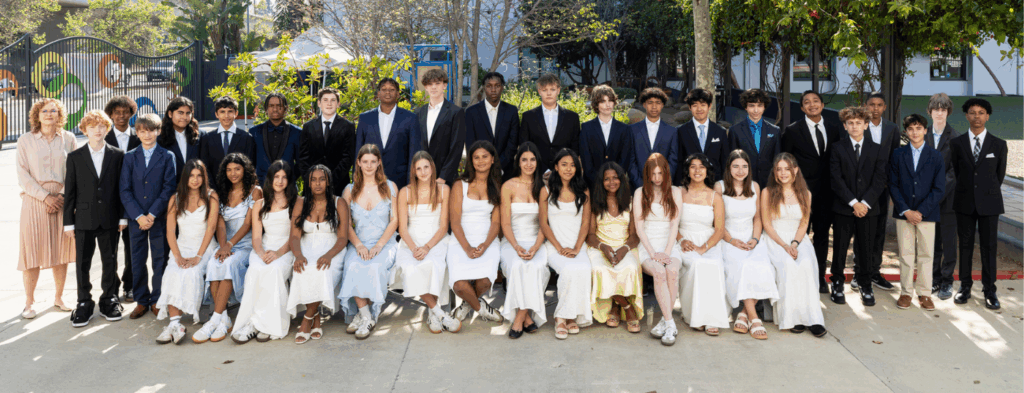Turning Point School’s 2025 Grade 8 graduating class posing outdoors in formal attire, with students arranged in two rows—seated in front and standing behind—alongside a faculty member. The group is diverse, smiling, and dressed in suits and light-colored dresses, with greenery and the school campus visible in the background.