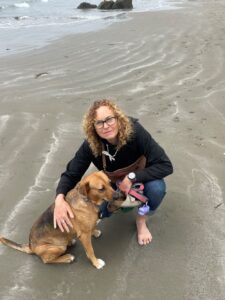 Woman smiling at the camera, kneeling next to her dog in the sand on a beach.